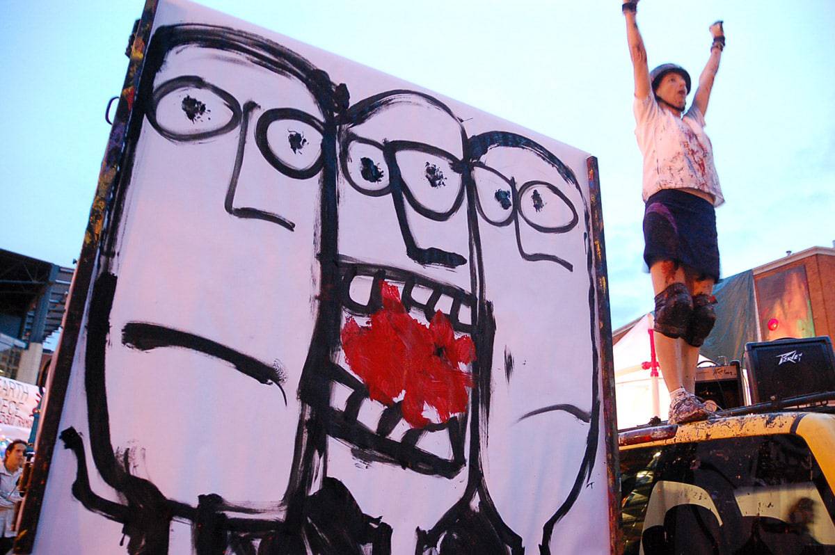 performance artist wearing helmet and kneepads stands on truck next to large painting of faces done in a modern primitive style by Julie JAO